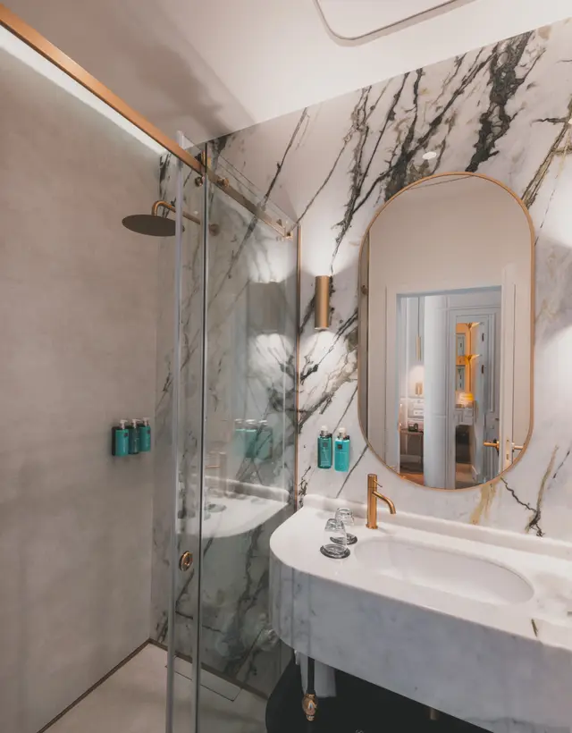 Baño de mármol con ducha de mampara, lavabo y espejo ovalado en una habitación interior de La Fonda de los Príncipes