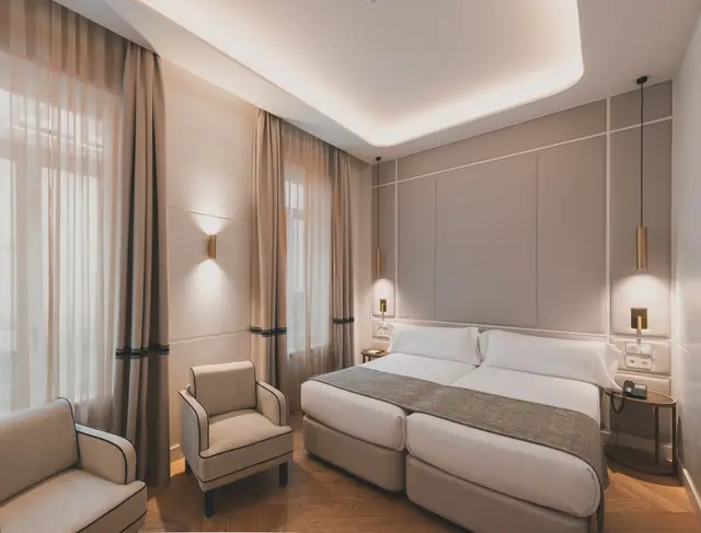 Camas con cabecero tapizado, apliques dorados y butaca junto al ventanal en la Deluxe Interior