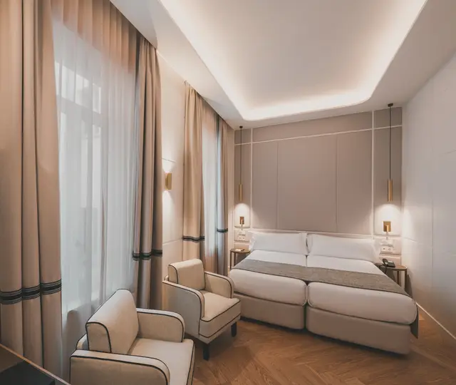 Habitación Deluxe Interior con dos camas, butacas y cortinas bajo una iluminación indirecta cálida