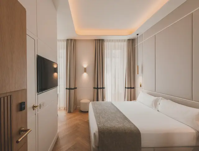 Cama doble con cabecero tapizado y cortinas junto a los ventanales de una habitación Deluxe Lateral Sol