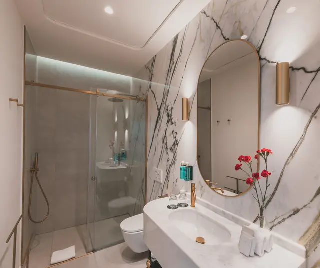 Lavabo de mármol, espejo ovalado de marco dorado y ducha con mampara en el baño de la Deluxe Vista Sol