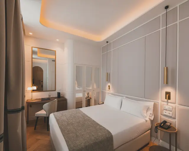 Cama con cabecero panelado y escritorio con espejo al fondo en la habitación Estándar Interior