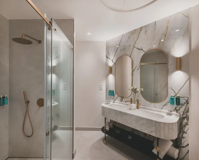 Baño de mármol con doble lavabo, espejos ovalados dorados y ducha de lluvia en la Premium Corner con bañera