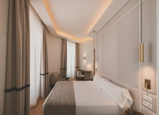 Cama doble, cortinas, butaca y escritorio al fondo de la habitación Premium Corner con bañera