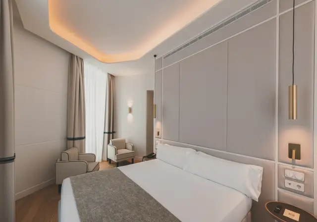 Cama doble, butacas y ventanal con cortinas al fondo de la habitación Premium Corner con ducha